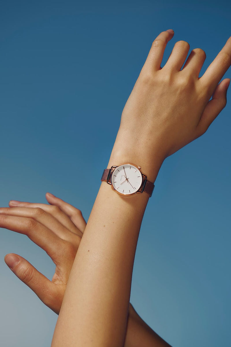 A minimalist Solios Curve Mini  solar watch with a white dial and rose gold case, worn on a person's wrist against a solid blue sky background.