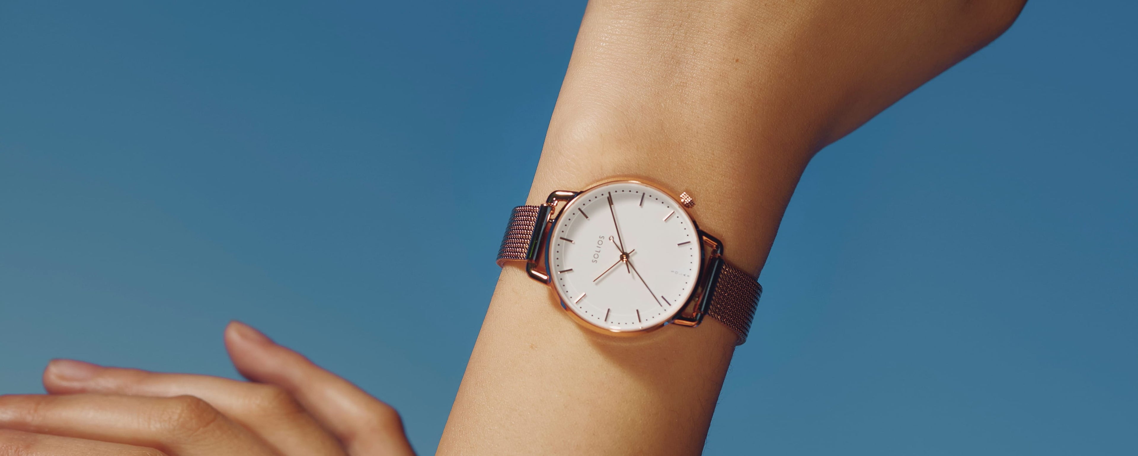 A close-up of a wrist wearing a minimalist Solar Curve Mini watch with a white dial and rose gold-toned case, paired with a brown leather strap, set against a solid blue background.