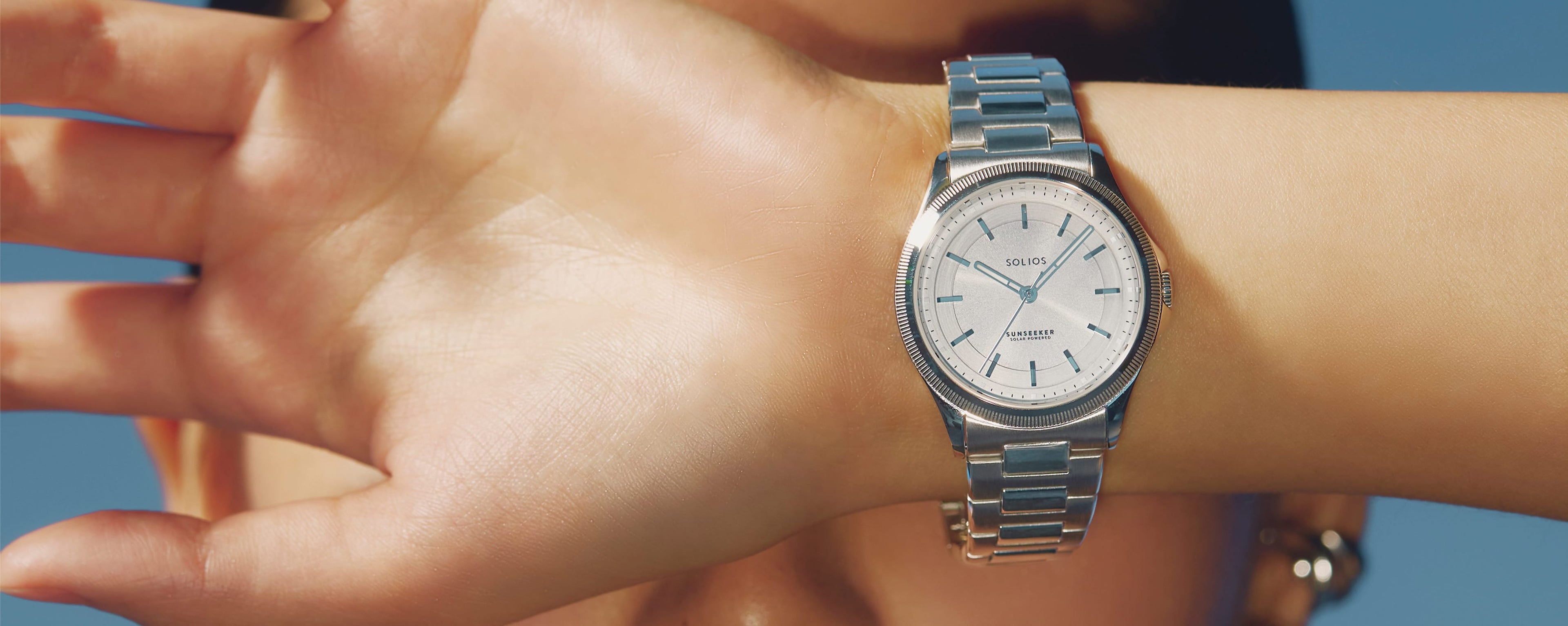 Close up of a women wrist wearing a silver dial, silver case Sunseeker solar watch, with a silver metal bracelet, hiding a woman's face in front of a blue sky.