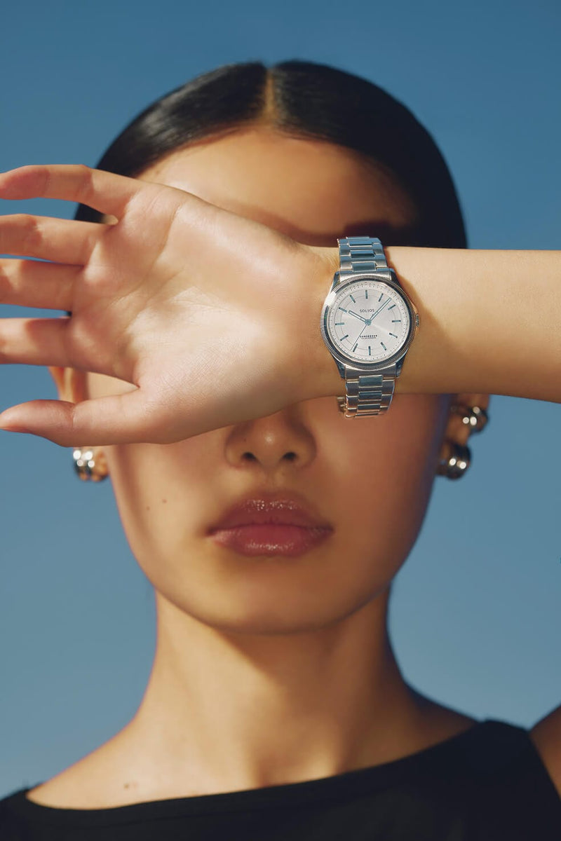 A silver-toned metal Sunseeker solar watch with a silver dial and silver link bracelet worn on a model's wrist, with her hand covering part of her face against a solid blue background.