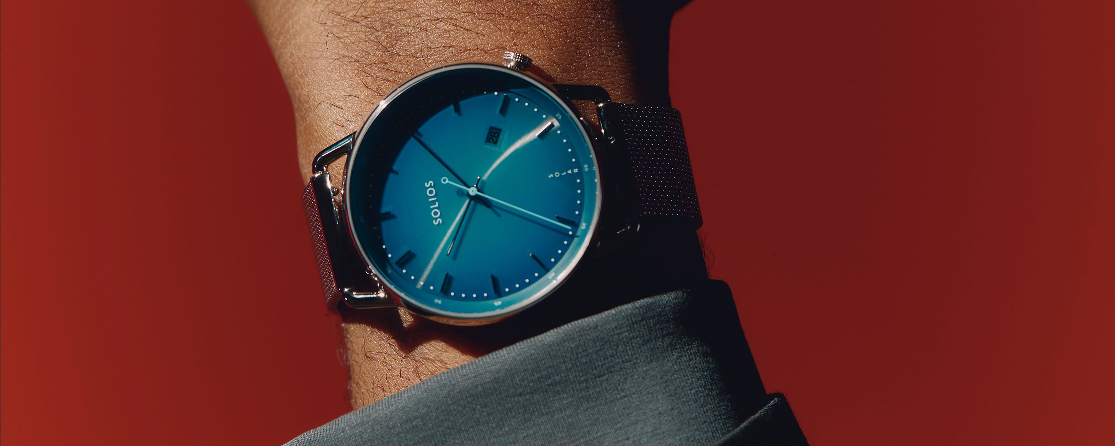Close up of a man's wrist wearing the Solar Curve with blue dial and silver case, on a silver mesh metal brand, in front of a red background