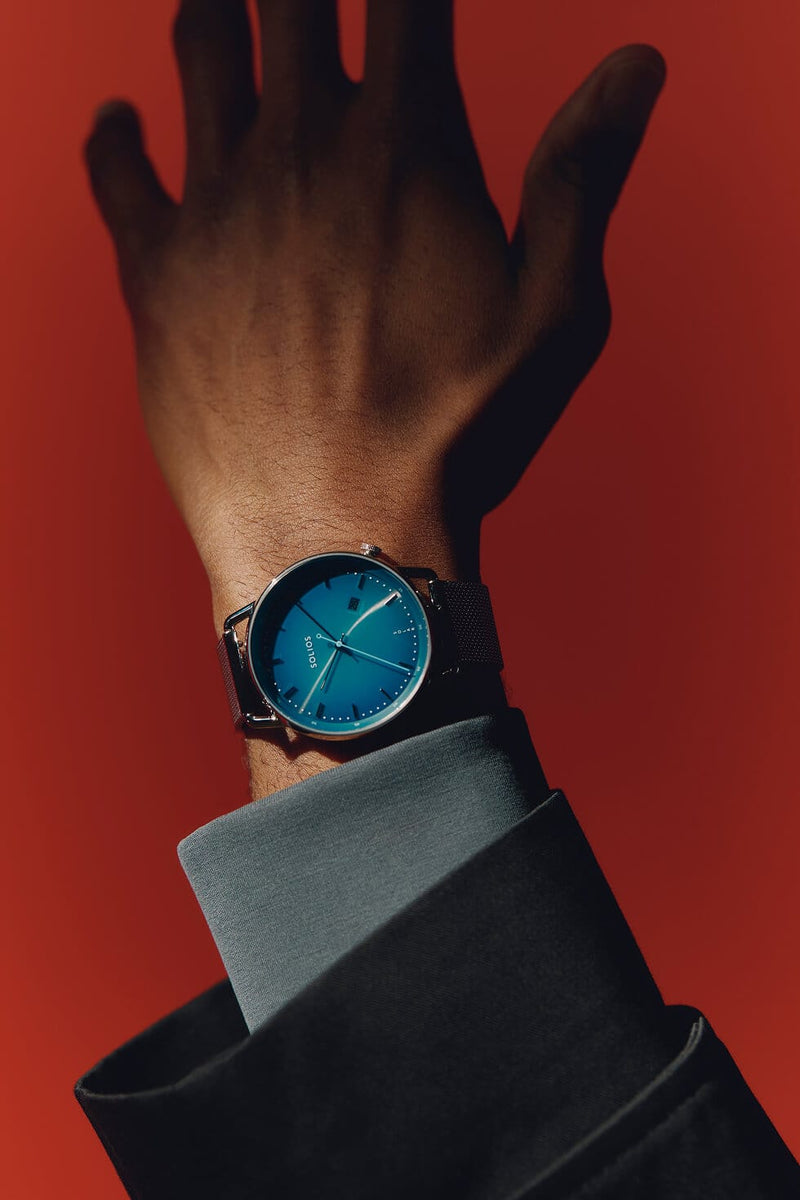 A Solios Curve solar watch with a blue dial and dark strap worn on a person's wrist, with the hand and dark suit sleeve visible against a solid red background.