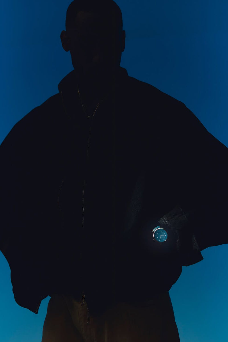 A dark silhouette of a person wearing a black jacket against a deep blue gradient background, wearing a glowing blue Solios Sunseeker watch on one wrist.