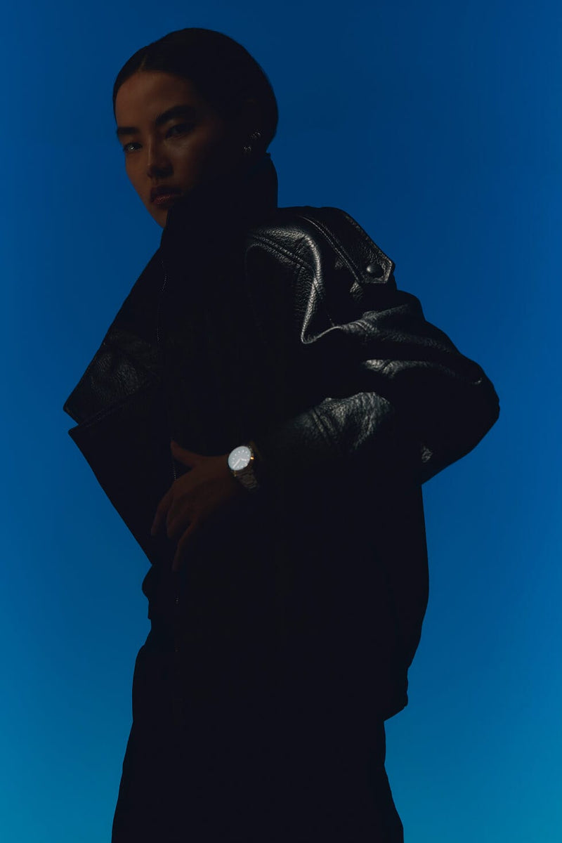 A model in a black leather jacket and black beanie poses against a solid blue background, wearing a silver-toned Solios solar watch on one wrist.