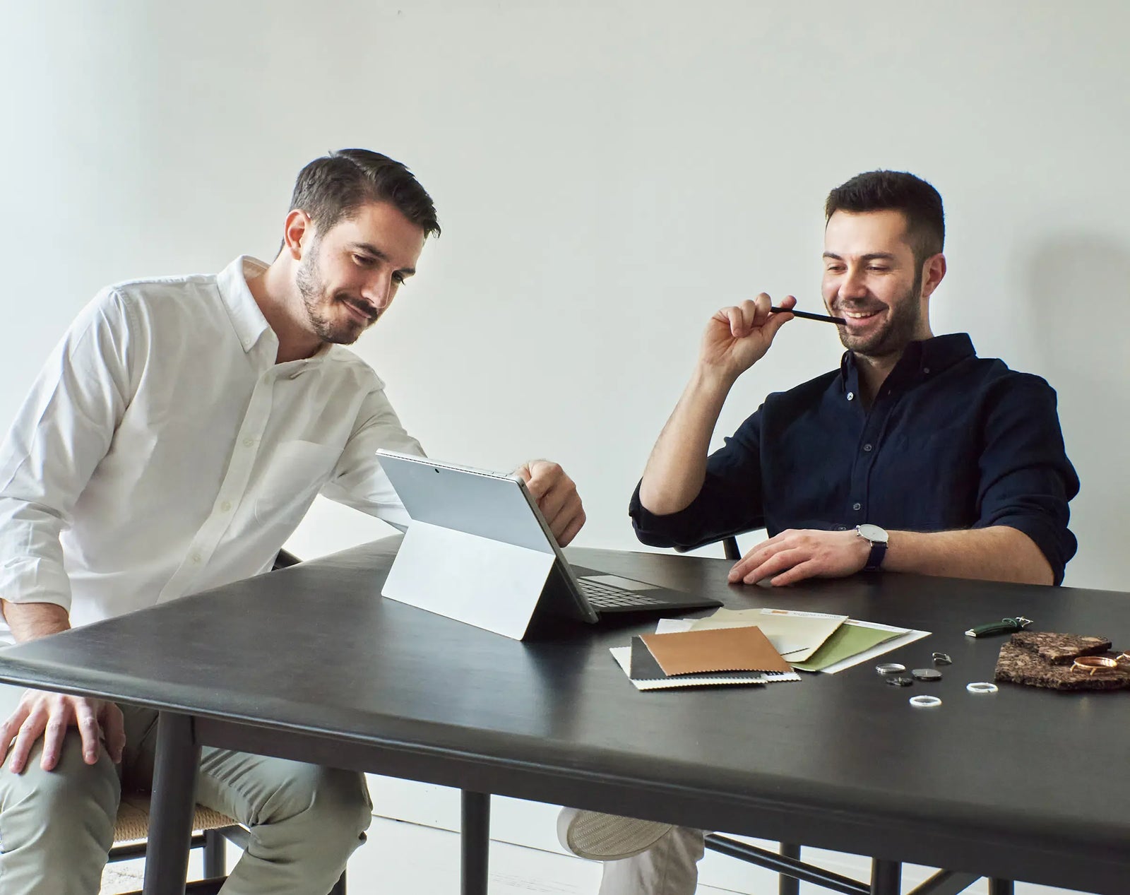 Sam and Alex, Solios cofounders, sitting at a dark table in a bright room, looking at a tablet and holding pens, designing solar watches, with papers and a small brown item on the table.