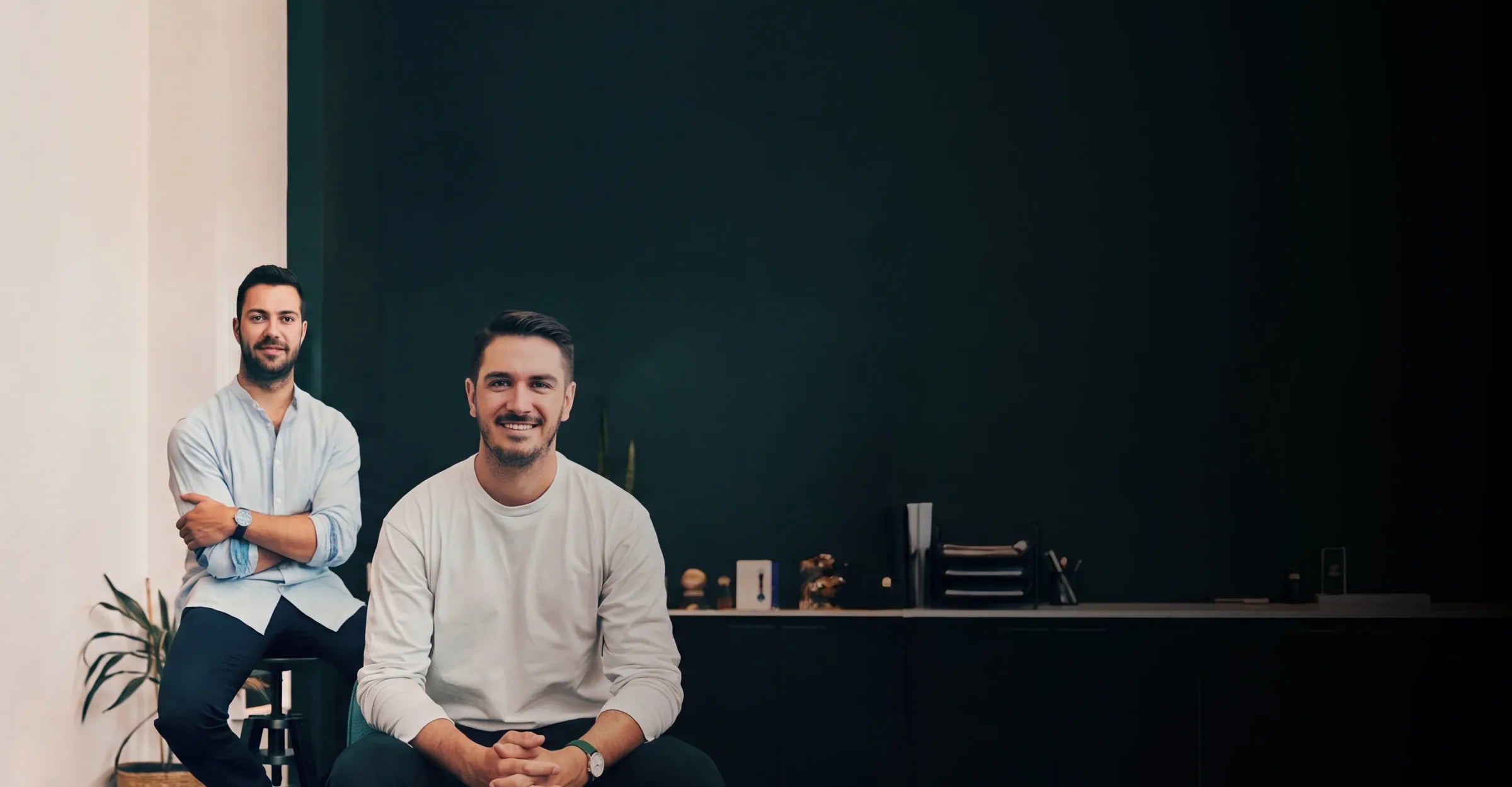 Sam and Alex, Solios cofounders, sitting in a modern office setting with a dark green wall behind them. The man in the foreground is wearing a light gray long-sleeve shirt, dark pants and a white Solios solar watch, while the man in the background wears a light blue button-down shirt, dark trousers and a blue Solios solar watch.