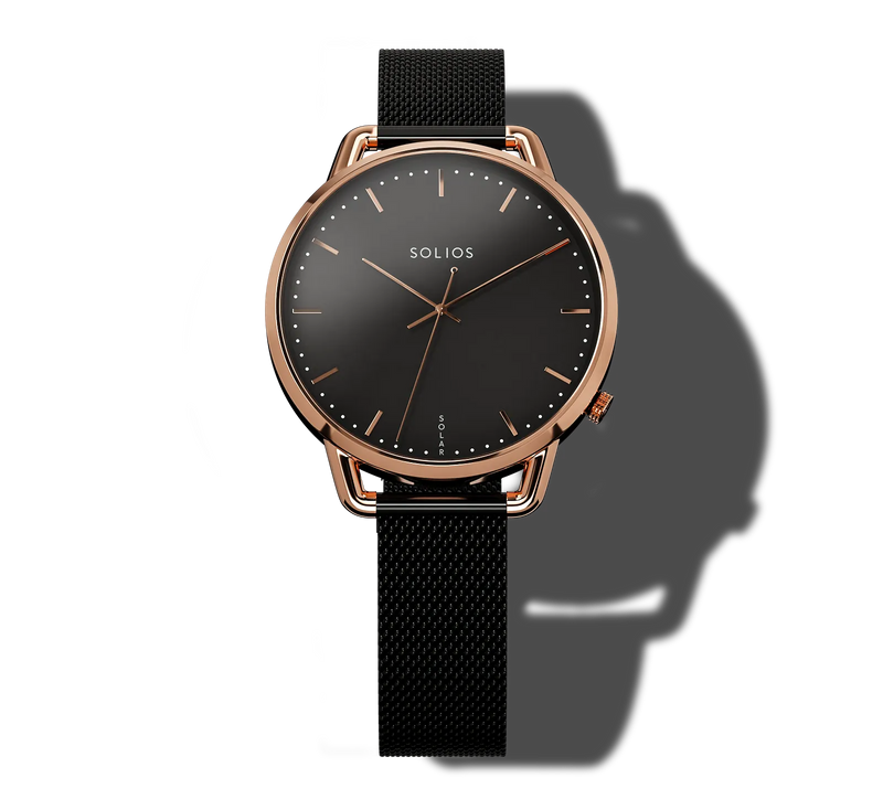 A Solios Curve Mini, round analog solar watch with a black dial, rose gold case, and black mesh strap, shown on a white background with a prominent shadow.