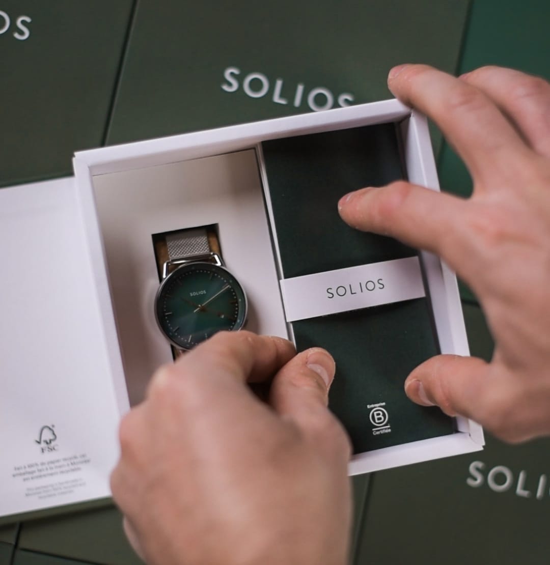 A SOLIOS watch with a dark green dial and a silver mesh strap is presented inside an open white Solios packaging, made from recycled and recyclable cardboard, in Montreal, with a person's hands holding the box open. The background consists of dark green SOLIOS branded packaging.