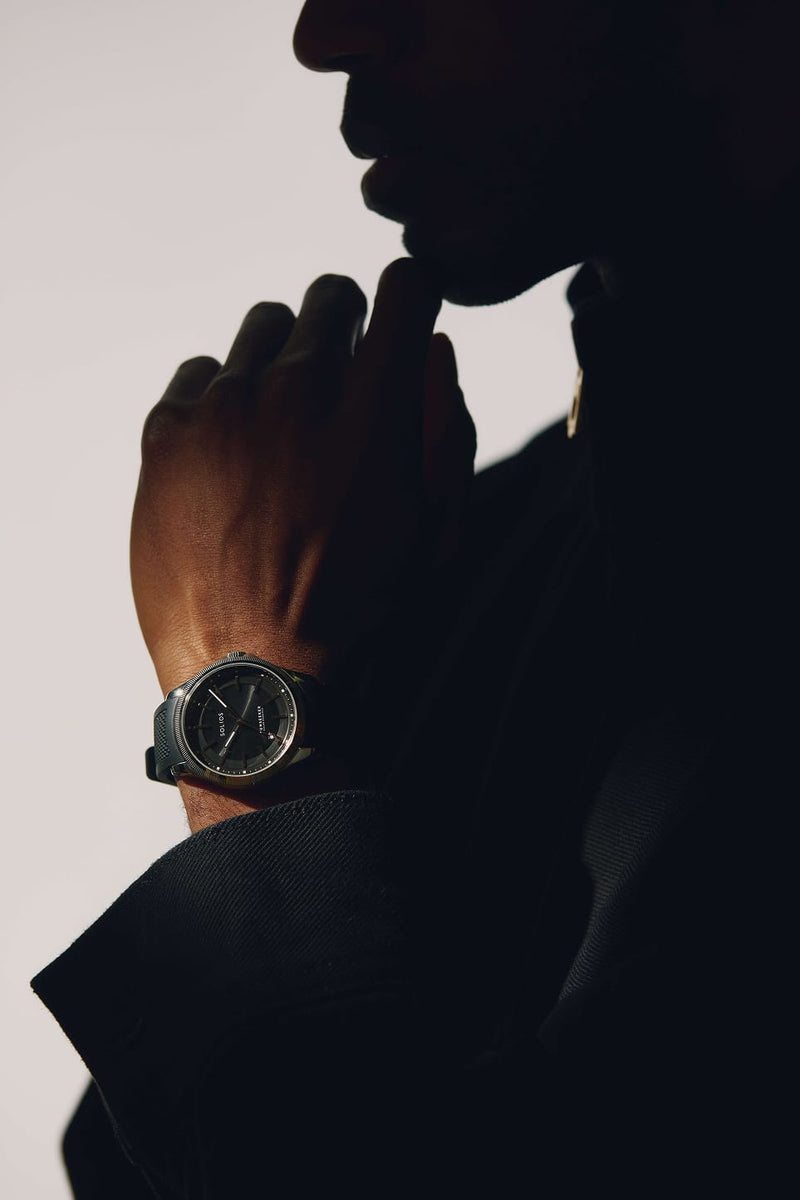 A close-up lifestyle shot of a man wearing a silver-toned Solios Sunseeker solar watch with a dark dial and strap, shown in dramatic low light against a light gray background.
