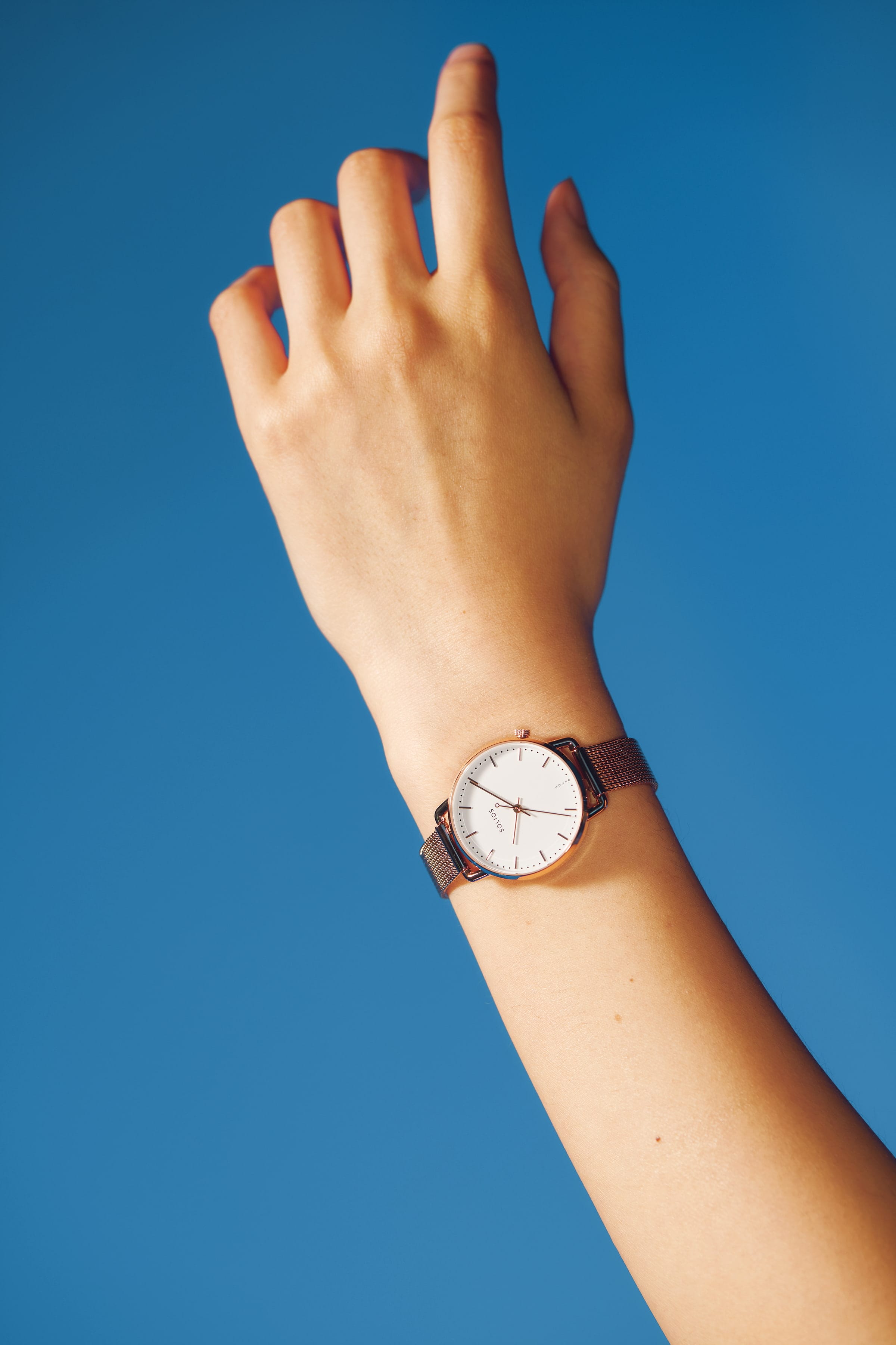 A minimalist Solios solar watch with a white dial and rose gold mesh strap worn on a person's wrist, set against a solid blue background.