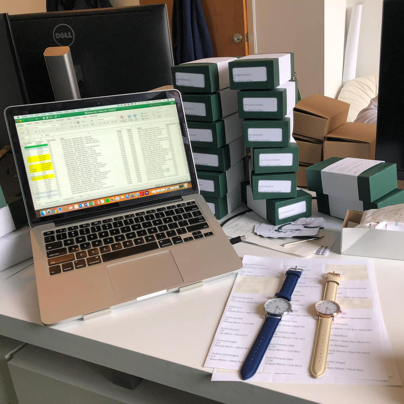 First two Solios watches on a desk beside a laptop and stacks of green shipping boxes announcing the delivery of the first Solios solar watches in April 2019.