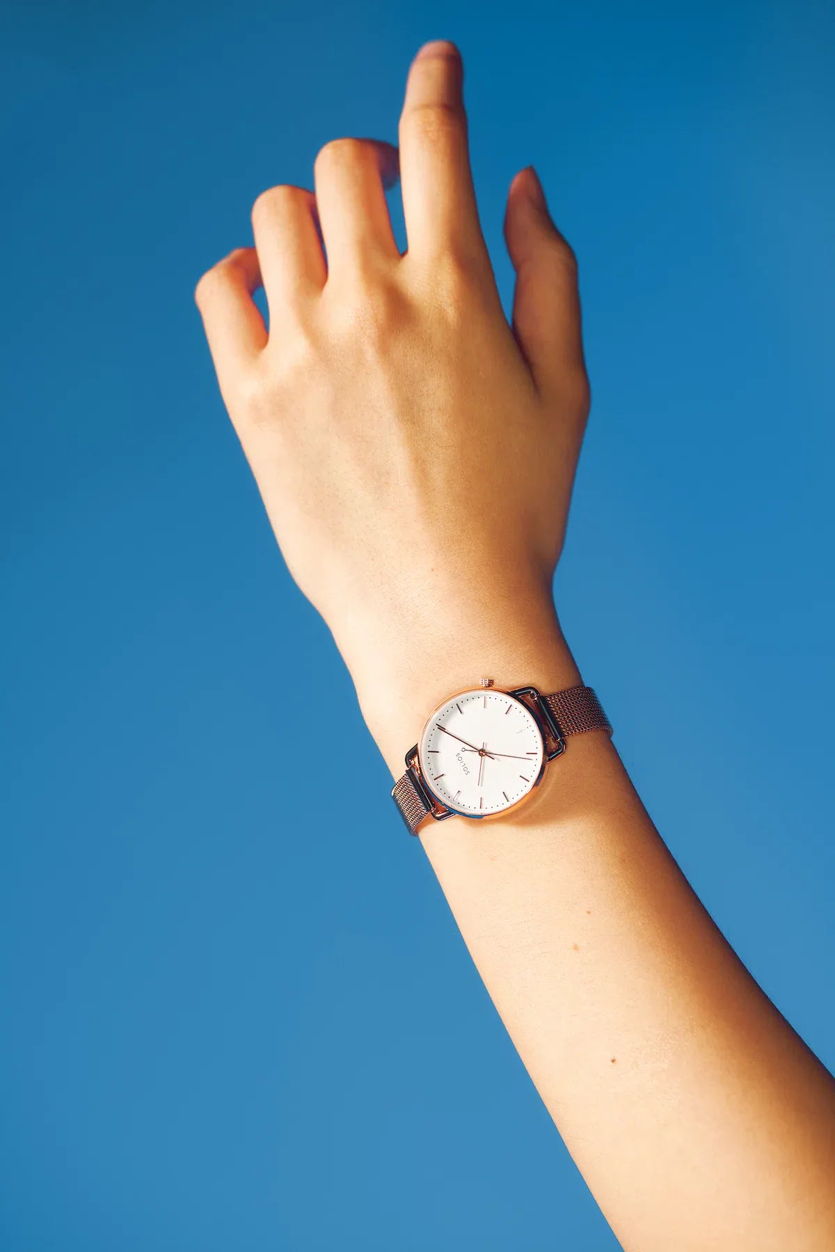 A minimalist Curve Mini solar watch with a white dial and brown leather strap worn on a person's wrist, set against a solid blue background.