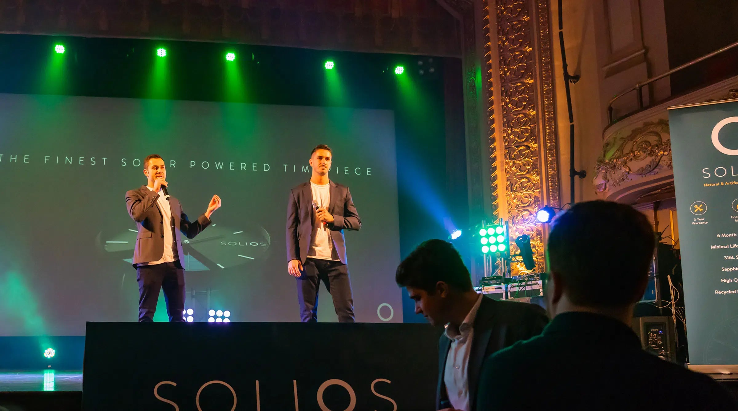 Solios founders presenting on stage at the 2018 Kickstarter launch night. We see a background banner saying "The finest solar powered timepiece".