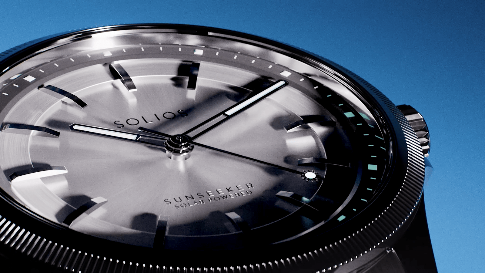 Close-up of a SOLIOS Sunseeker solar watch with a silver dial, black bezel, and black case against a clear blue sky.