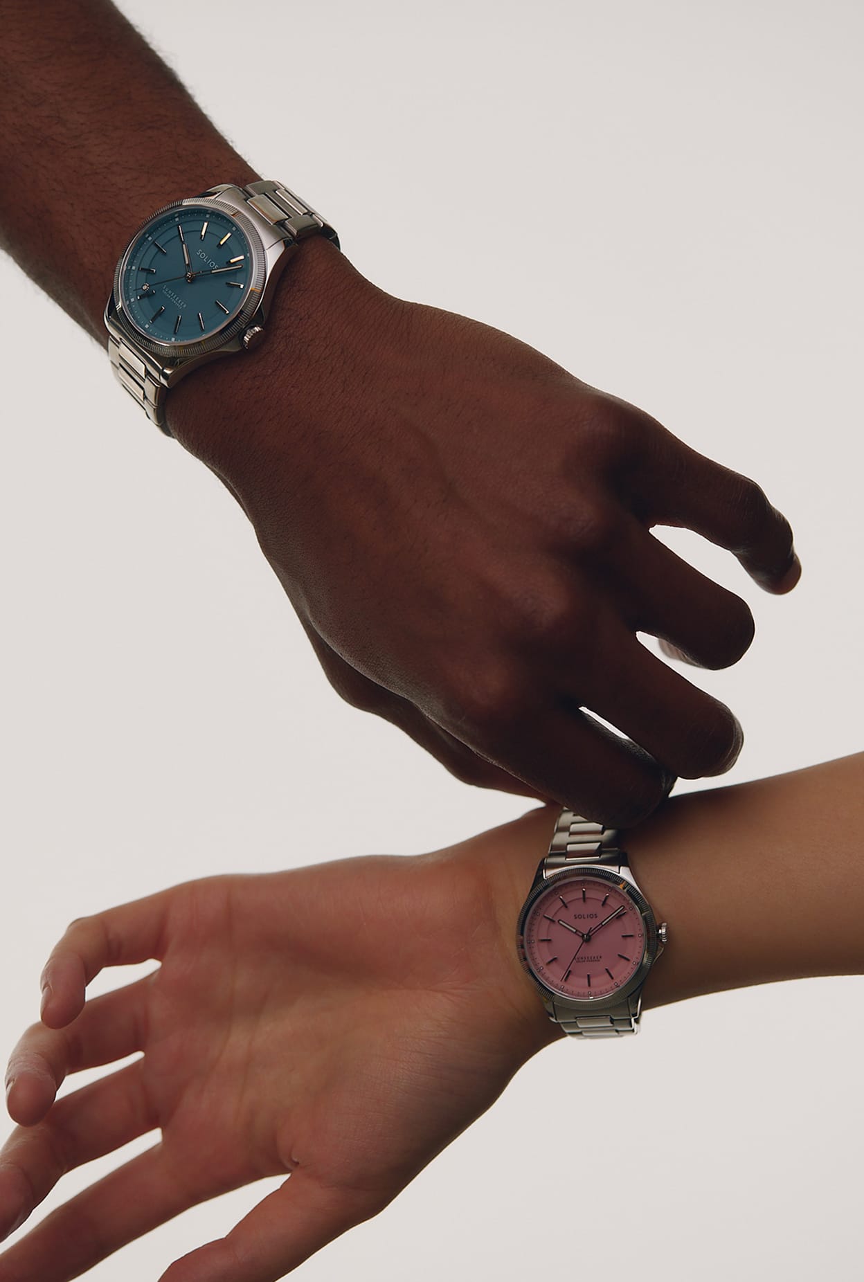 Two Solios Sunseeker analog solar wristwatches with silver metal bracelets and round dials, one with a blue face and the other with a pink face, shown worn on two different hands against a plain light background.
