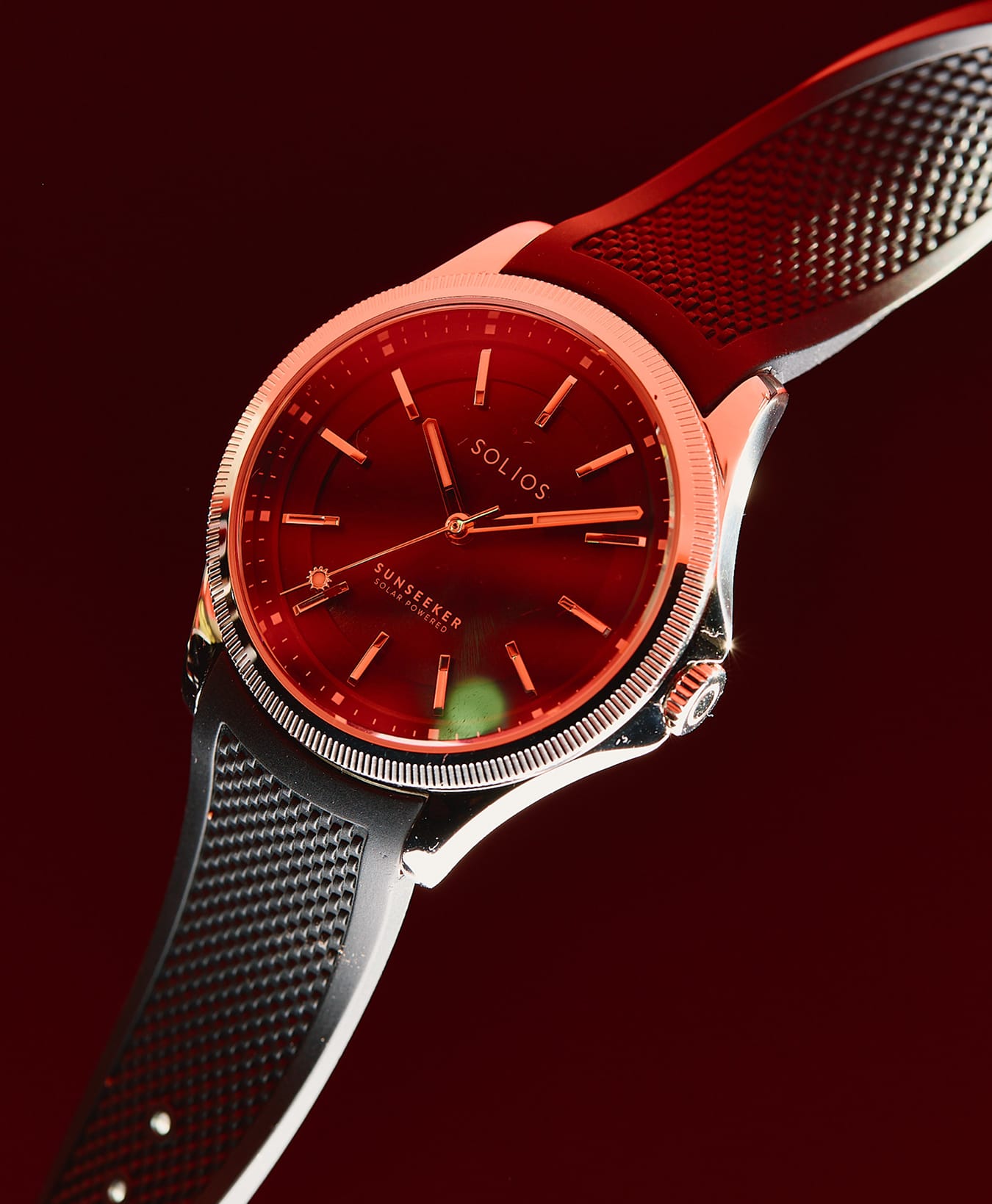 A close-up of a SOLIOS Sunseeker solar watch with a deep black dial, silver-toned case, and a textured black rubber strap, set against a dark red background create a red reflection on the watch.